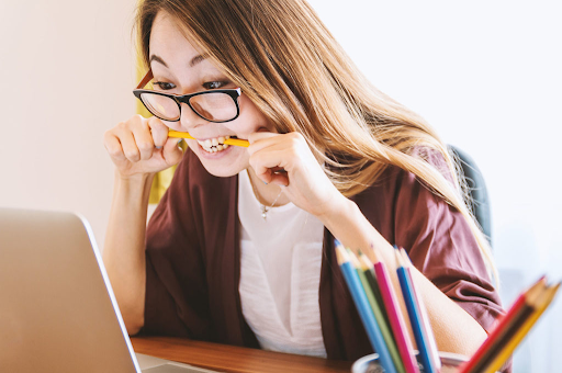 The image shows a woman wearing glasses, biting a pencil, and looking at a laptop screen with a stressed expression. On her desk are colorful pencils in a holder.