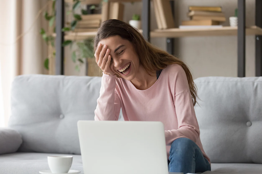 The image shows a woman sitting on a couch, laughing while looking at her laptop. She has her hand on her forehead, indicating she's enjoying something funny on the screen.