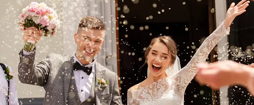 The image shows a joyful bride and groom exiting a building, possibly after their wedding ceremony. They are smiling and celebrating, with the groom holding a bouquet and guests throwing confetti.