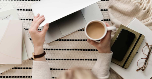 The image depicts a person’s hands—one holding a cup of coffee and the other interacting with a laptop. The setting suggests a work or study environment